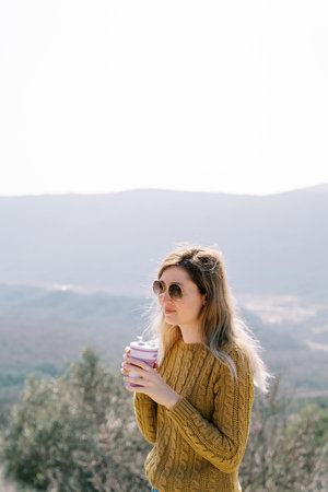 Smiling female traveler with a glass of coffee stands on a mountain