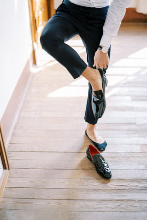 Groom puts on black patent leather shoes while leaning on the wall. Cropped. Facelessの写真素材