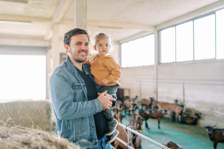 Smiling dad with a little girl in his arms stands at the pen with goatsの写真素材