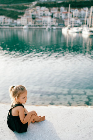 Little girl sits half-turned on the pier and looks away. Back viewの写真素材