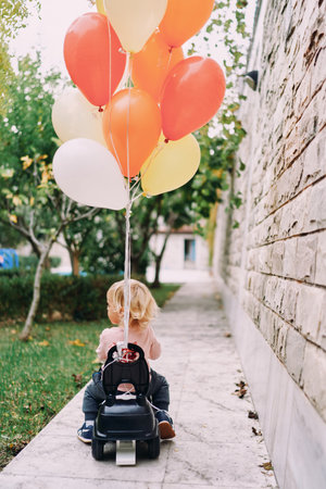Little girl is driving a toy car with tied balloons around the yard. Back viewの写真素材