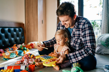 Dad plays with a little girl on a colorful xylophone, sitting behind her on the bedの写真素材