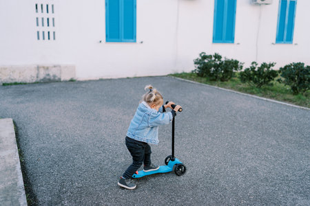 Little girl rides a scooter on an asphalt yard near the house