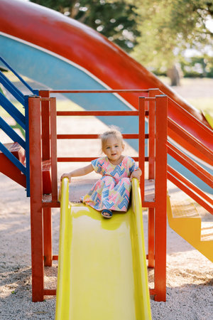 Little girl sits on top of a slide on the playground holding on to the railingsの写真素材