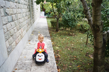 Little girl rides a colorful toy car along the wall in the gardenの写真素材