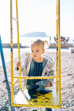 Little girl stands with a soft toy near the swing, holding on to the handrailsの写真素材