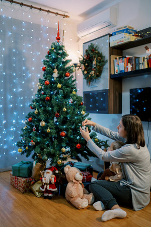 Little girl watches her mom decorate the Christmas tree with balls. Side viewの写真素材