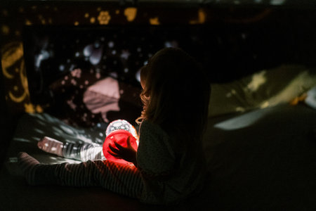 Little girl sits on the bed with a night-light projector in her hands and looks at the pictures on the wallsの写真素材
