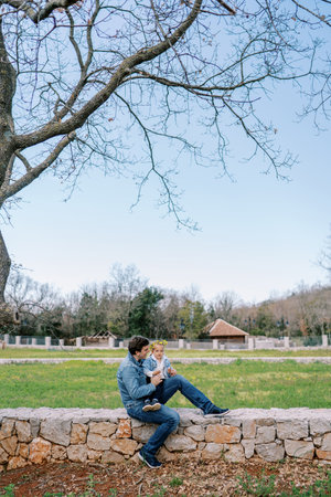 Dad with a little girl in a wreath on his knees sits on a stone fence in a flowering meadowの写真素材