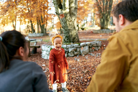 Little laughing girl under falling leaves stands in front of mom and dad in the autumn park. Back viewの写真素材