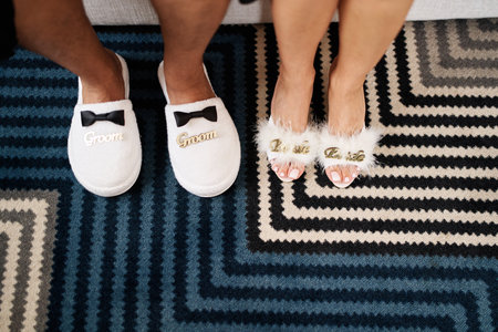 Male and female feet in slippers stand on a striped carpet. Caption: Groom. Bride. Cropped, faceless