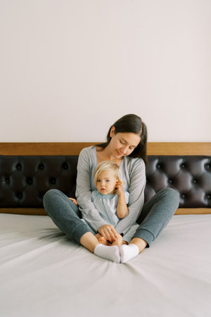 Mom hugs a little girl sitting behind her on the bedの写真素材