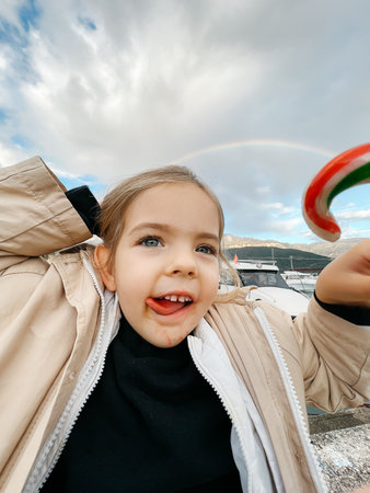 Little girl with her tongue hanging out and a candy in her hand against the backdrop of a rainbowの写真素材