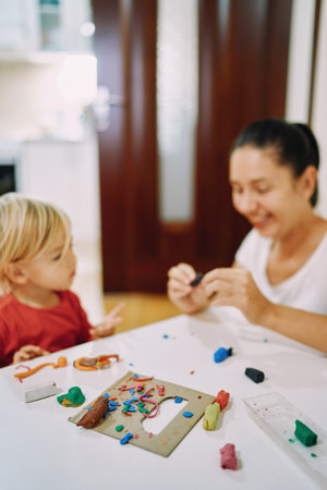 Mom and her little daughter sculpt different figures from colored plasticine while sitting at the...