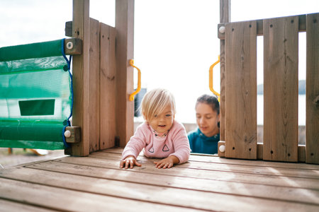 Little girl crawls on her stomach on a wooden deck of a slide against the background of her motherの写真素材
