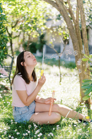 Young girl sits under a tree on the lawn and blows soap bubbles
