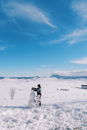 Mom and little girl are making a snowman, standing on a snowy clearing in a mountain valley. Back viewの写真素材