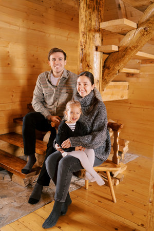 Smiling dad sitting on the stairs next to mom and little girl sitting on a chair in a wooden cottageの写真素材