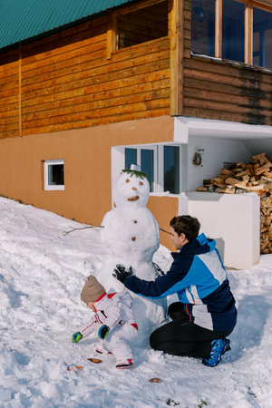 Dad and little girl decorate a snowman with buttons near a woodpile of a wooden chalet in the snow. Back viewの写真素材