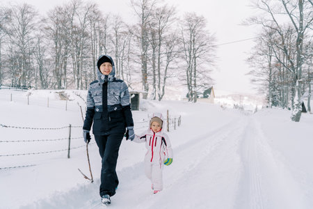 Mother and a little girl walk holding hands along a snowy road on the edge of a forestの写真素材