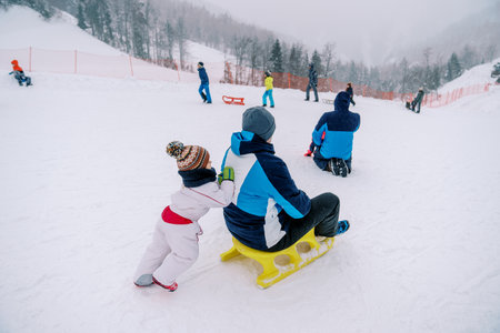 Small child pushes his dad on a sled down a snowy hill. Back viewの写真素材
