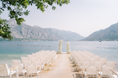 Rows of white chairs stand in front of a wedding arch on a pier by the sea against the backdrop of mountainsの写真素材