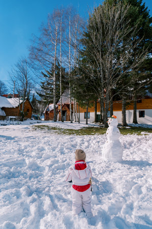 Little girl stands next to a snowman in the courtyard of a wooden house. Back viewの写真素材