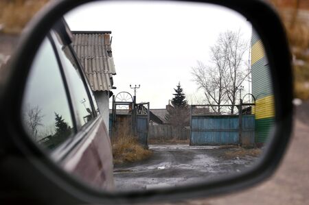 Reflection in car mirror: the open gate, the old roof of the building, dirt road, tree, poplar. Early spring.の写真素材