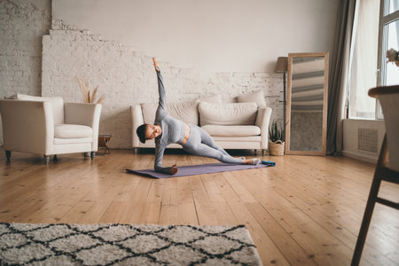 Doing sports at home online. Sports girl doing exercises. A girl in a gray tracksuit goes in for sports. Young woman exercising at home in a living room.の写真素材
