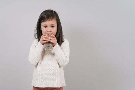 The child drinks water from a glass. A girl with dark hair holds a glass of water. The brunette leads a healthy lifestyleの写真素材