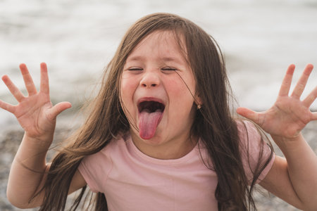 The child indulges and pranks. A little girl shows her tongue. Funny girl writhes her face. Little girl wants to become an actressの写真素材