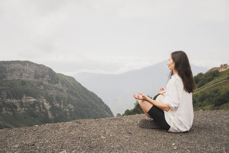A girl meditates in the mountains. Yoga Retreat in Nature. A girl in a white shirt enjoys the beautiful nature. The girl sits in the lotus position with her eyes closed.の写真素材