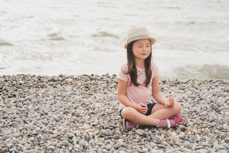 A conscious girl meditates on the shore by the sea. A little girl does yoga. Psychological maturation of modern children. A girl in a pink T-shirt dreamsの写真素材