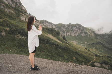 A girl meditates in the mountains. Yoga Retreat in Nature. A girl in a white shirt enjoys the beautiful nature. The girl sits in the lotus position with her eyes closed.の写真素材