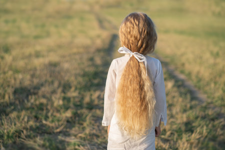 A child with beautiful curly long hair. A little girl in a white dress stands with her back. A beautiful ribbon in the hair of a child. Girl in a simple dress in the fieldの写真素材