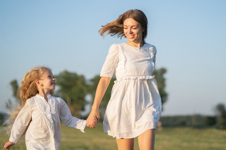 A mother and daughter run into the field by the hand. Happy childhood with my mother. Daughter and mother in a white dress in the field. Psychology of parents relations with childrenの写真素材