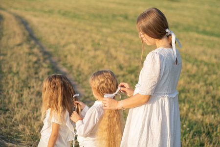 A gentle mother with two daughters in nature. Mom braids beautiful hair for her daughters. Care for beautiful and thick hair of a blonde. Psychology of the relationship between mother and daughter.の写真素材