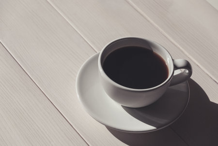 Black coffee in white cup with a saucer on light wooden table, shadow, copy space