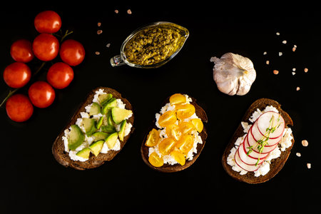 Set of vegan sandwiches with cheese, avocado, radish, yellow cherry tomatoes and herbs on a black background. Pesto in a gravy boat, red cherry tomatoes on branch, garlic head. Top view.の写真素材