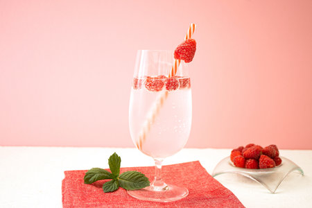 One glass of refreshing lemonade with raspberries stands on a red napkin on a white table against a...