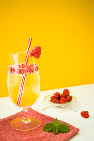 A summer soft drink with raspberries in a glass stands on a red textile napkin and on a white table on an yellow background. Near is a bowl of sweet raspberries and fresh mint leaves. Verticallyの写真素材