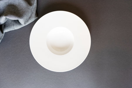 An empty white round plate stands on a gray background, next to it lies a gray towel. Stylish table...