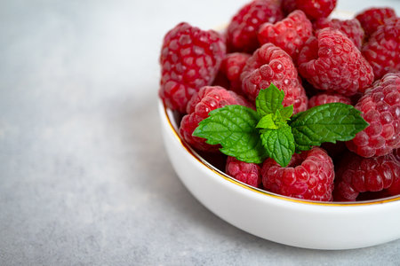 Sweet ripe raspberries with mint leaves in a white bowl with a gold rim stands on a gray concrete background. Healthy dessert, proper nutrition. Seasonal fresh berries. Close-upの写真素材