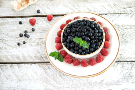 Delicious blueberries in a white bowl sit on a white spacer with a gold rim on a light wooden table. The counter plate is decorated with sweet raspberries and mint leaves. Nice presentation. Top viewの写真素材