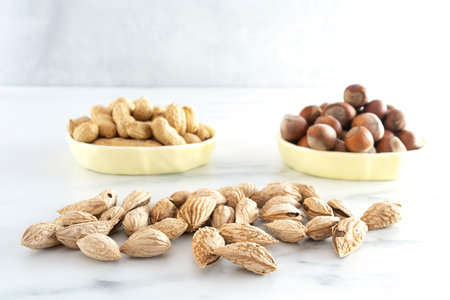 A handful of in-shell almonds lie on a light table. In the background are two bowls of hazelnuts and...