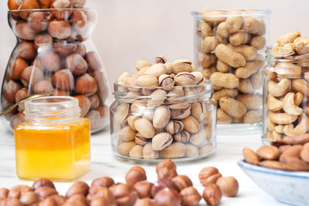 Pistachios in a glass jar stand on a white table. Nearby are jars of honey and nuts (hazelnuts in...