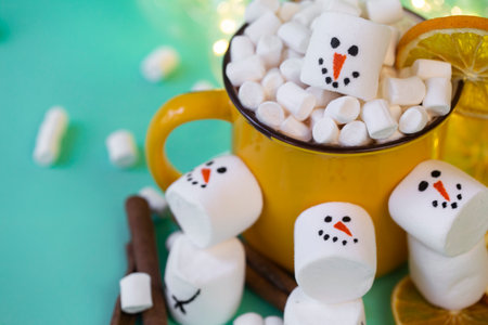 marshmallow snowmen on a mint turquoise background with a yellow cocoa mug illuminated by a garland close upの写真素材