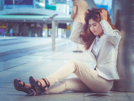 A young asian businesswoman is confused and stressed , plucking her head and sitting outside office. Unemployment or economy concept.の写真素材