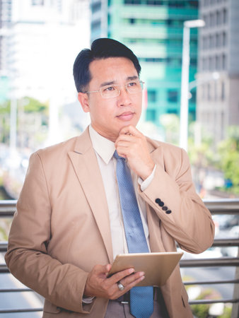 The asian business man in a suit is pondering and holding digital tablet outside corporate office.の写真素材