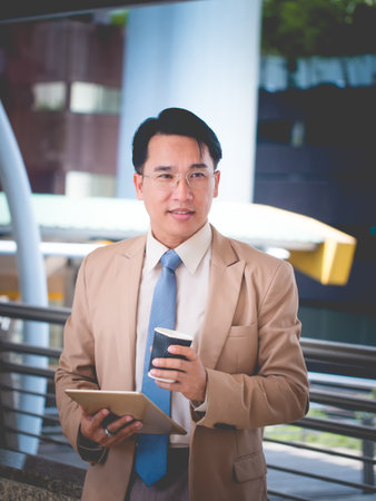 The smart asian businessman in a suit and wearing glasses holding his digital tablet and a cup of coffee outside corporate office.の写真素材